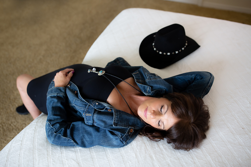 Older lady in a sexy black dress and denim jacket leaning against the bed.  Photo by Spark Boudoir