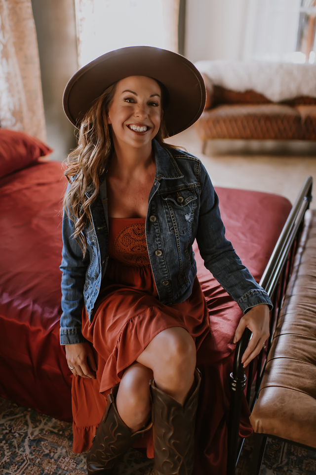 Kellie Foster in a brown hat, orange dress and jean jacket with cowboy boots taking a photo for Spark Boudoir. 